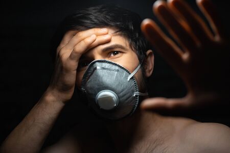 Virus concept. Dramatic dark portrait of young man wearing protective flu mask, holding head with...