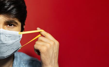 Studio close-up portrait of young guy, half face, puts on medical flu mask, protection from coronavirus, isolated on red background with copy space.の写真素材