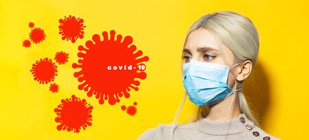 Portrait of young blonde girl with blue eyes, looking at coronavirus icon and covid-19 text, on background of yellow color.の写真素材