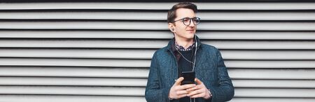 Panoramic banner, portrait of young man wearing eyeglasses, holding smartphone listening music with...