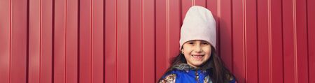 Panoramic portrait of smiling child girl on background of red metal outdoor wall.の写真素材
