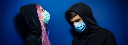 Dark studio portrait of young hooded couple, guy and girl wearing medical flu mask prevention of coronavirus on blue background.の写真素材
