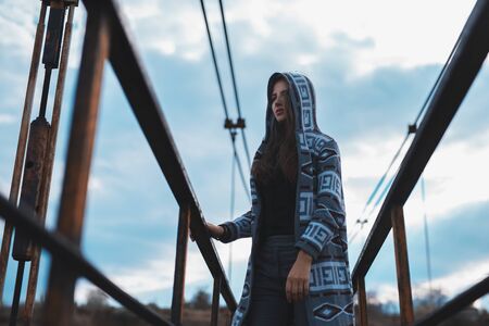 Dramatic outdoor portrait of young teenage hooded girl standing on abandoned old rusty bridge with perspective background.の写真素材