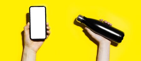 Close-up of male hands holding smartphone with mockup and reusable steel thermo water bottle isolated on yellow background.の写真素材