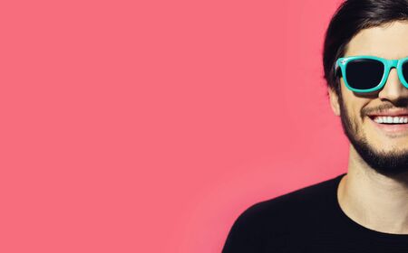 Studio half face portrait of young smiling guy wearing cyan sunglasses on background of pastel pink...