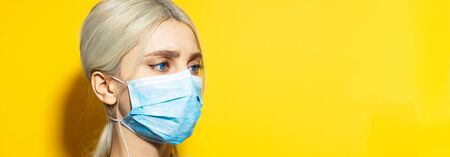 Panoramic portrait of young blonde girl with blue eyes, wearing medical face mask against coronavirus on yellow background with copy space.の写真素材