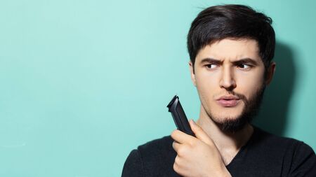 Portrait of young guy with half shaved face holding trimmer in hand on background of aqua menthe color with copy space.の写真素材