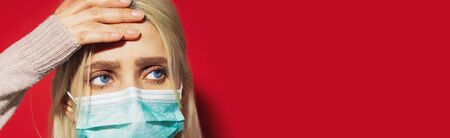 Portrait of young blonde girl with blue eyes, having headache, holding hand on head, wearing medical face mask against coronavirus on red background with copy space.の写真素材