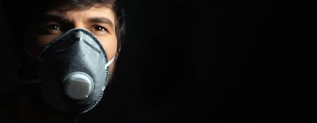 Coronavirus prevention. Close-up portrait of young guy wearing protective medical respiratory mask against flu, on black background with copy space.の写真素材