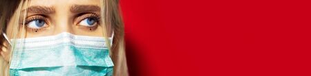 Close-up of young female face with blue eyes, wearing respiratory medical face mask against coronavirus on red background with copy space.の写真素材