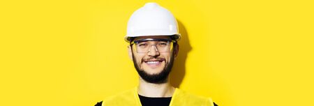 Studio panoramic portrait of young smiling construction engineer worker man, wearing safety helmet and goggles on background of yellow color with copy space.の写真素材