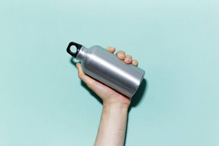 Close-up of male hand holding reusable, aluminum thermo bottle for water, on studio background of cyan, aqua menthe color. Zero waste. Plastic free.の写真素材