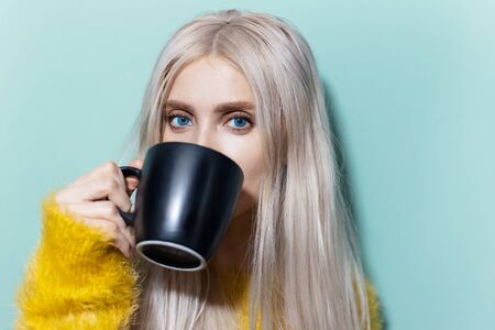 Studio portrait of beautiful blonde girl with blue eyes drinking tea from black cup, wearing yellow sweater on cyan, aqua menthe color.の写真素材