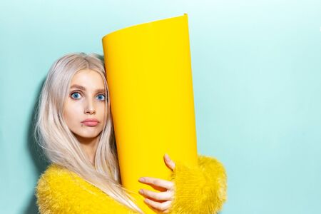 Studio portrait of young excited girl holding big yellow roll of paper in hands. Background of cyan,...