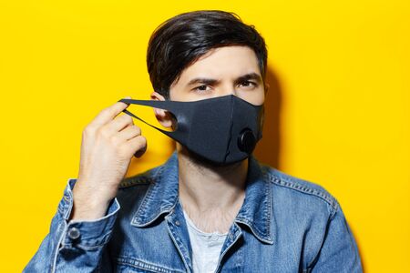Studio portrait of young guy, putting on black respiratory face mask against coronavirus, background...