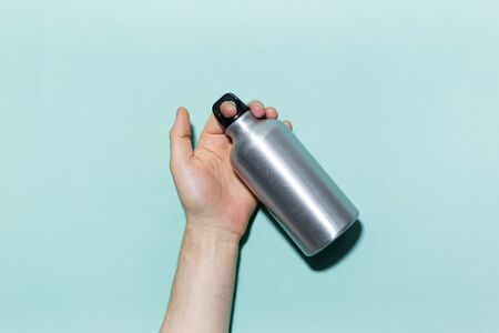 Close-up of male hand holding eco, reusable aluminum thermo water bottle on studio background of cyan, aqua menthe color.の写真素材