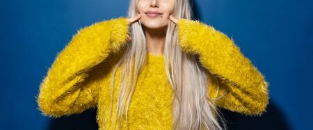 Young girl in yellow sweater pointing fingers on cheeks, on blue background.