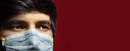 Close-up of male face wearing medical flu mask against coronavirus on dark red background with copy...