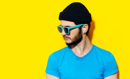 Studio portrait of young hipster guy using wireless earphones, wearing blue shirt, cyan sunglasses and black beanie hat on background of yellow color with copy space.の写真素材