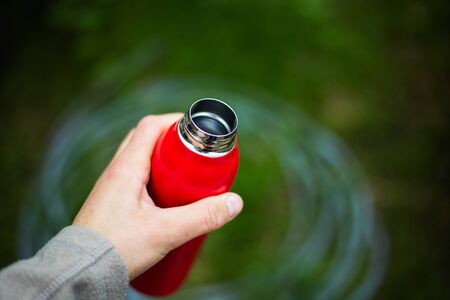 Close-up of male hand holding red reusable steel thermo water bottle on background of green bush in bokeh.の写真素材