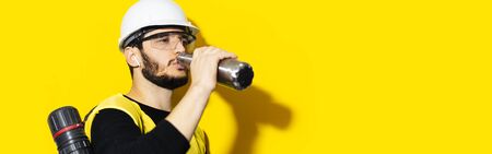 Studio portrait of young engineer man wearing safety helmet, goggles and jacket. Using wireless earphones, drinking water from steel thermo reusable bottle. Background of yellow with copy space.の写真素材