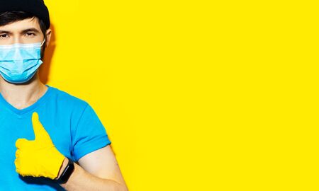 Panoramic banner, portrait of young guy showing thumbs up. Wearing smartwatch, safety gloves and medical face mask, protection against coronavirus. Covid-19 prevention. Studio background of yellow.の写真素材
