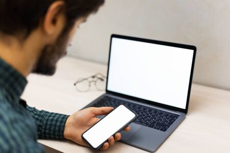 Back view close-up of young guy holding modern smartphone with mockup beside laptop on work desk.の写真素材