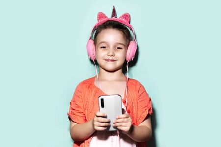 Colorful portrait of happy confident little child girl, holding smartphone and listening the music with headphones on studio background of cyan, aqua menthe color.の写真素材