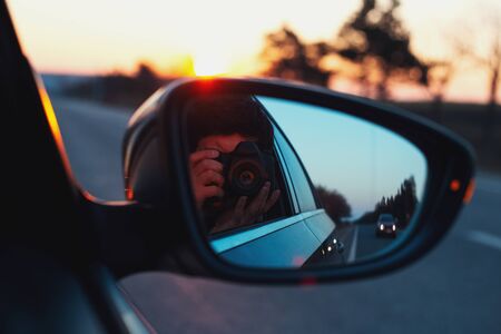 Reflection in car mirror of young guy taking photo of sunset with digital camera.の写真素材