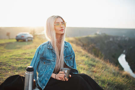 Portrait of young blonde girl on camping, wearing denim jacket and yellow glasses, holding steel cup with tea near. Background of sunset.の写真素材