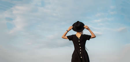 Back view of young blonde girl, wearing black dress and hat, on background of sky.の写真素材