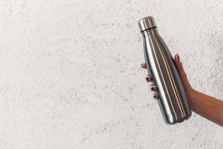 Close-up of female hand holding reusable steel thermo water bottle on background of textured wall.の写真素材
