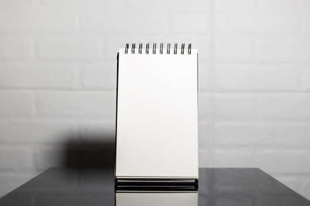 Close-up of empty page of notebook on black glass table, background of white brick wall.