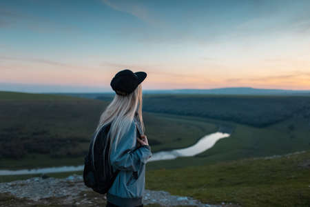 Back view of young blonde girl with backpack enjoying sunset on peak of hills. Travel concept.の写真素材