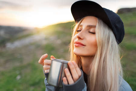 Close-up portrait of young blonde girl with black cap, holding steel mug with hot tea on background...