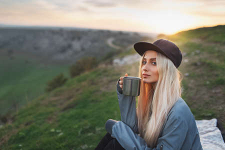 Portrait of young blonde girl with black cap, holding steel mug with hot tea on background of sunset.の写真素材
