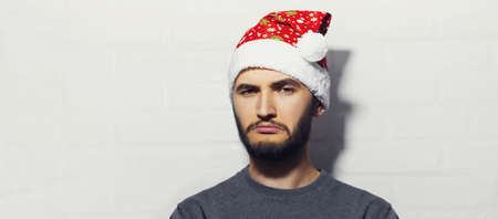 Panoramic portrait of young confident man wearing Santa Hat on background of white brick wall with copy space.の写真素材