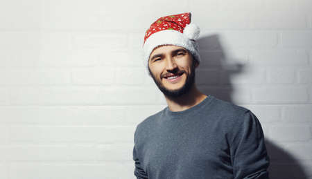 Studio portrait of young smiling guy on background of white brick wall. Christmas concept. Wearing red Santa Hat and grey sweater.の写真素材