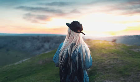 Back view of young blonde girl with black cap and backpack, walking on peak of the hills. Background of sunset.の写真素材