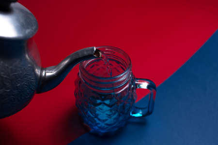 Close-up of aluminum teapot pouring water in glass mug for juice, on two backgrounds of red and blue colors. Top view.の写真素材