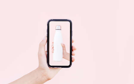 Close-up of female hand holding thermo bottle on screen of smartphone in another hand, on background...