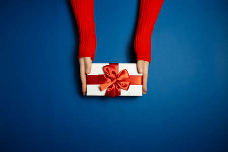 Top view of female hands holding gift box with red bow on background of phantom blue of color with copy space. Wearing sweater of lush lava color.の写真素材