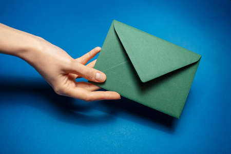 Close-up of female hand holding a green paper envelope on background of blue color.