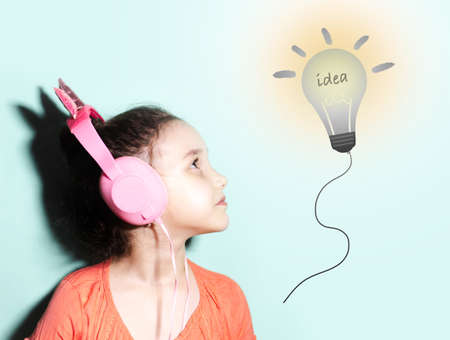 Studio portrait of child girl with headphones looking at light bulb; having new idea. Cyan...