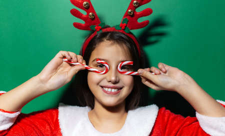 Studio portrait of happy child girl with striped candy on eyes. Wearing reindeer horns and Santa costume.の写真素材