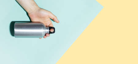 Close-up of male hand holding reusable thermo bottle. Backgrounds of pastel blue and yellow colors. Panoramic photo with copy space.の写真素材