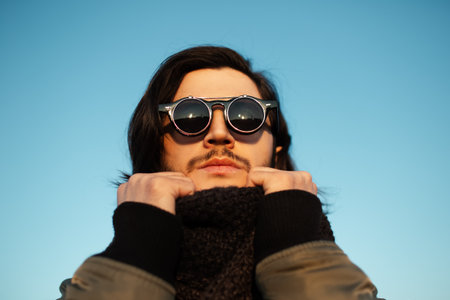 Close-up portrait of young guy with long hair wearing hipster round sunglasses and scarf. Blue sky on background.の写真素材