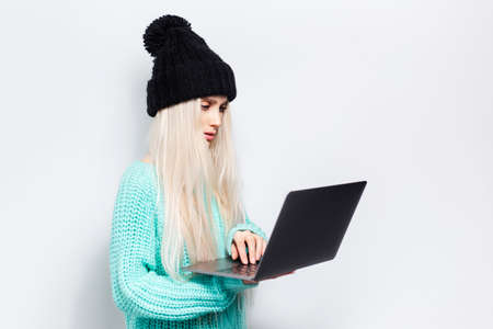 Studio portrait of young blonde girl using laptop on white background. Wearing black hat and cyan...