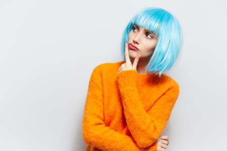 Studio portrait of thoughtful young girl, holding hand under chin against white background with copy space. Wearing orange sweater and blue wig.の写真素材