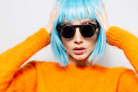 Close-up portrait of young fashionable confident girl on white studio background. Woman with blue hair wearing round sunglasses and orange sweater.の写真素材
